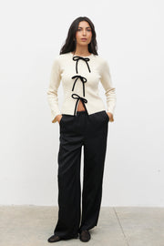 Woman wearing a white sweater with black ribbons and black pants against a white background