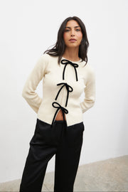 Woman wearing a cream cardigan with black ribbons and black pants against a white background