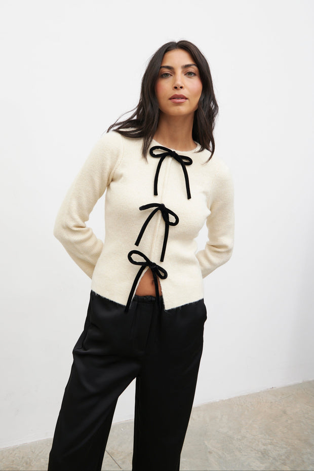 Woman wearing a cream cardigan with black ribbons and black pants against a white background